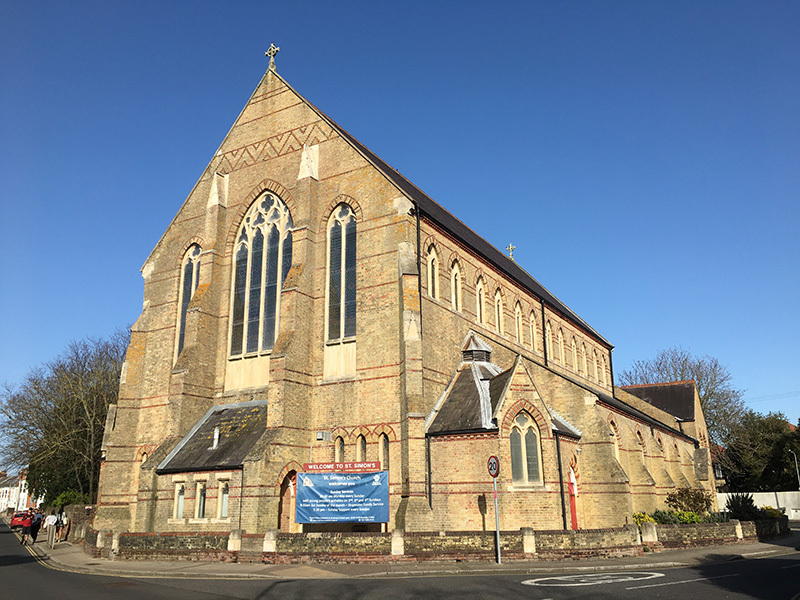 Exterior view of the west and southern faces of St Simon’s Church, viewed from the junction of Waverley Road and St Ronan’s Road. The church is brightly lit by late afternoon spring sunshine against a cloudless blue sky.