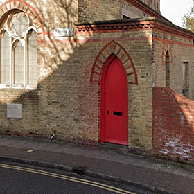 The red door of St Simon’s church office set into the north end of the east wall running along St Ronan’s Road.