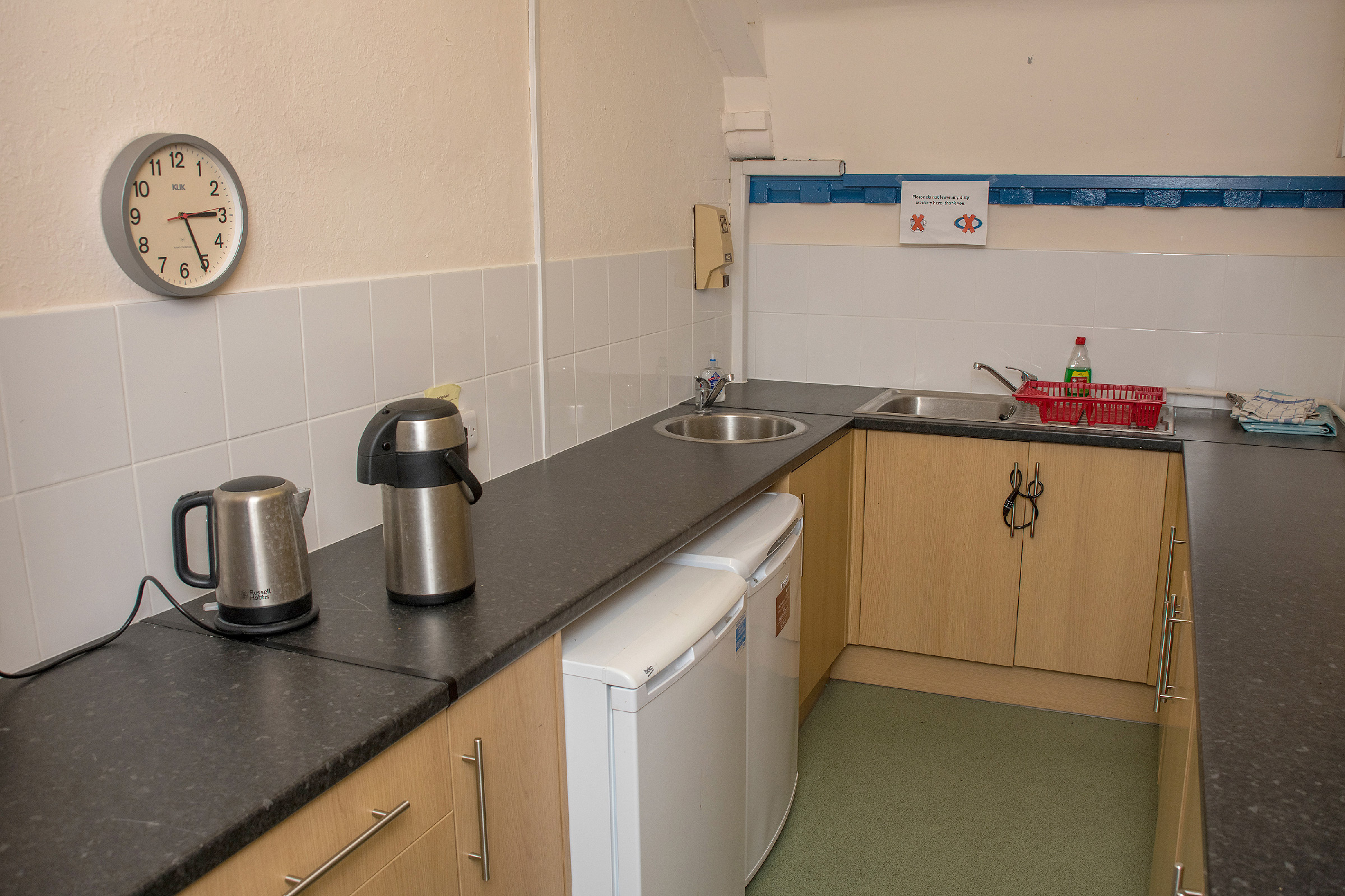 The kitchen adjacent to the upper hall with its dual sinks set into a dark worktop atop 6 cupboards. A kettle and thermos flask sit on the worktop to one side.